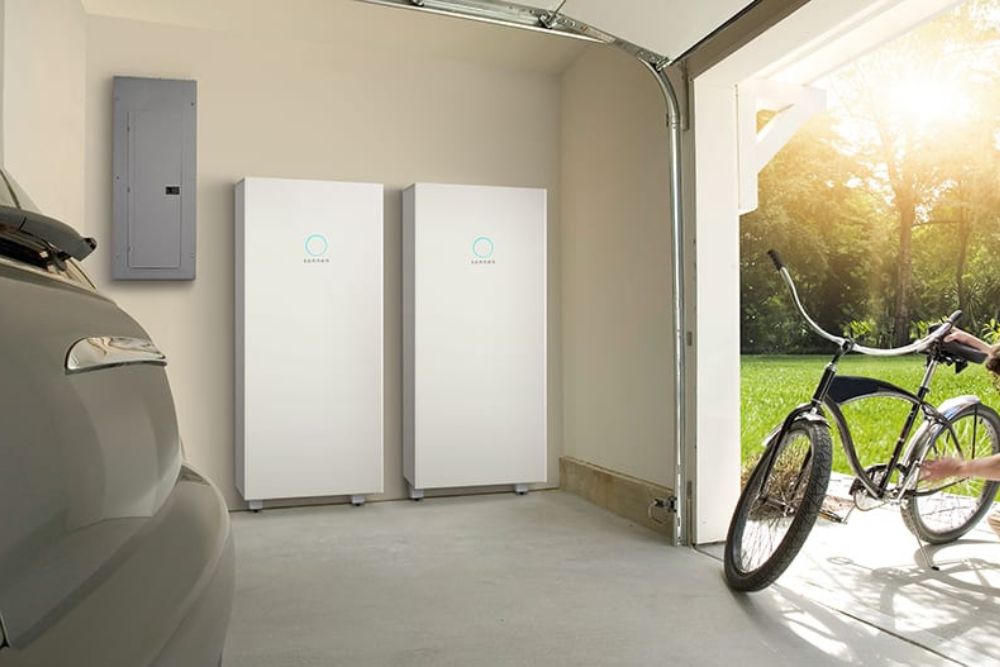 Two solar batteries mounted on a garage wall that has its door open to allow a bike and car to go in and out.